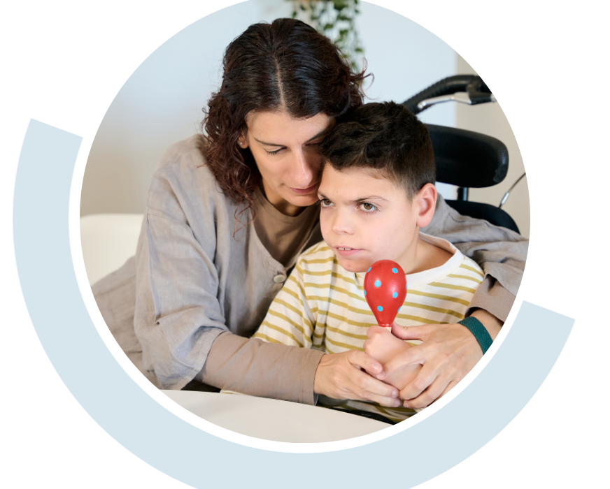 Website header graphic with a circular photo framed by a pale blue curved accent, featuring a woman closely supporting a seated young boy. She gently guides his hands as he holds a red maraca during a shared activity.