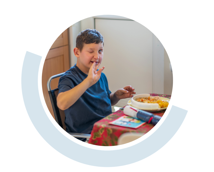 Website header graphic with curved light blue accents, featuring a circular photo of a young man seated at a dining table with a plate of food in front of him, smiling and gesturing with one hand.
