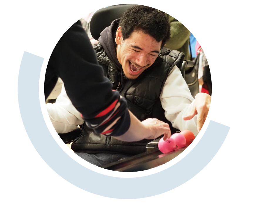 Website header graphic with a circular photo framed by a pale blue curved accent, featuring a smiling young man seated in a wheelchair and laughing during an interactive activity. Another person reaches towards colourful pink and red balls positioned in front of him.