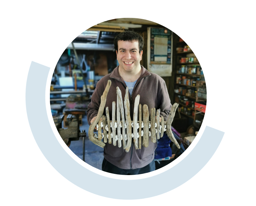 Website header graphic with a pale background and curved light blue accents, featuring a circular photo of a smiling young man standing behind a display of handmade wooden items in a workshop.