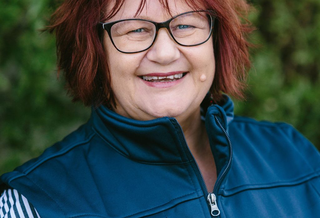 Head and shoulders portrait of a smiling woman with short reddish-brown hair and glasses standing outdoors in front of greenery, wearing a DIsability Connect vest.
