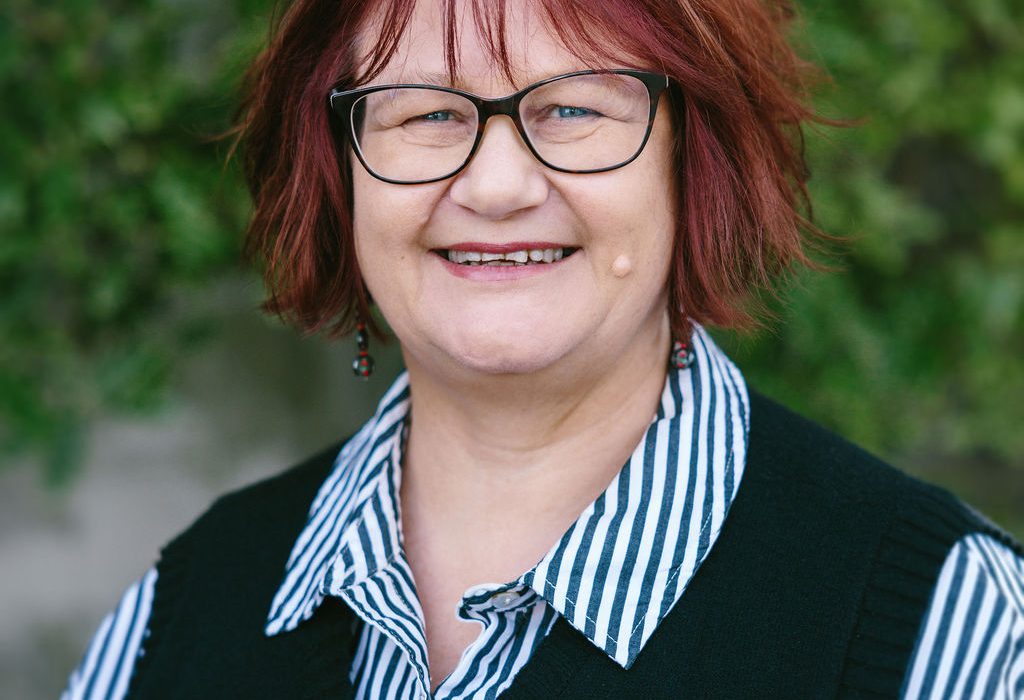 Head and shoulders portrait of a smiling woman with short reddish-brown hair and glasses standing outdoors in front of greenery.