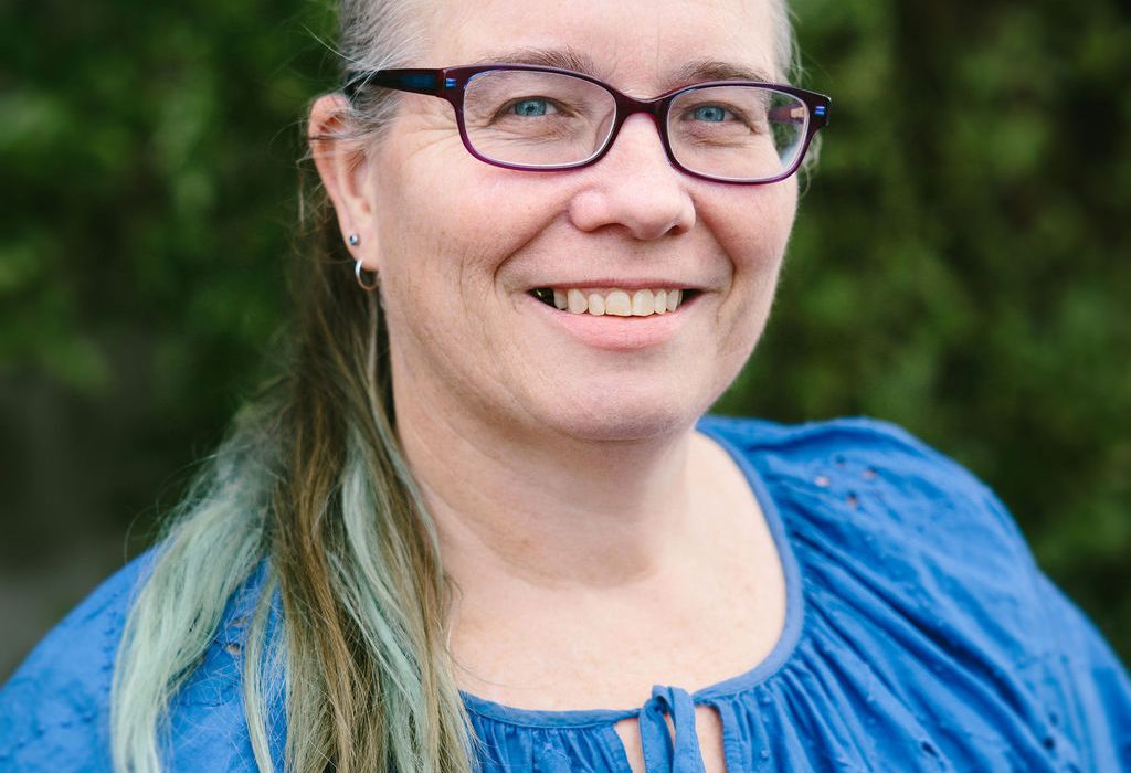 Head-and-shoulders portrait showing a smiling woman with her light brown, gray and blue hair pulled back, wearing purple rectangular glasses and a blue top against a leafy green outdoor background.