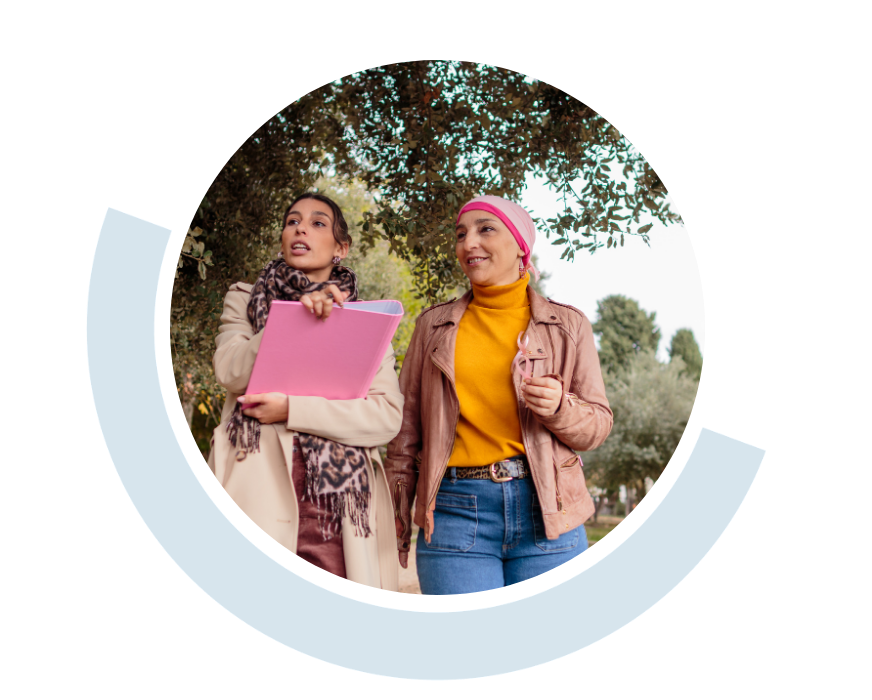 Website header graphic with a pale background and curved light blue accents, featuring a circular photo of two women walking and talking together outdoors, one holding a folder or document, the other wearing a pink scarf covering her head.
