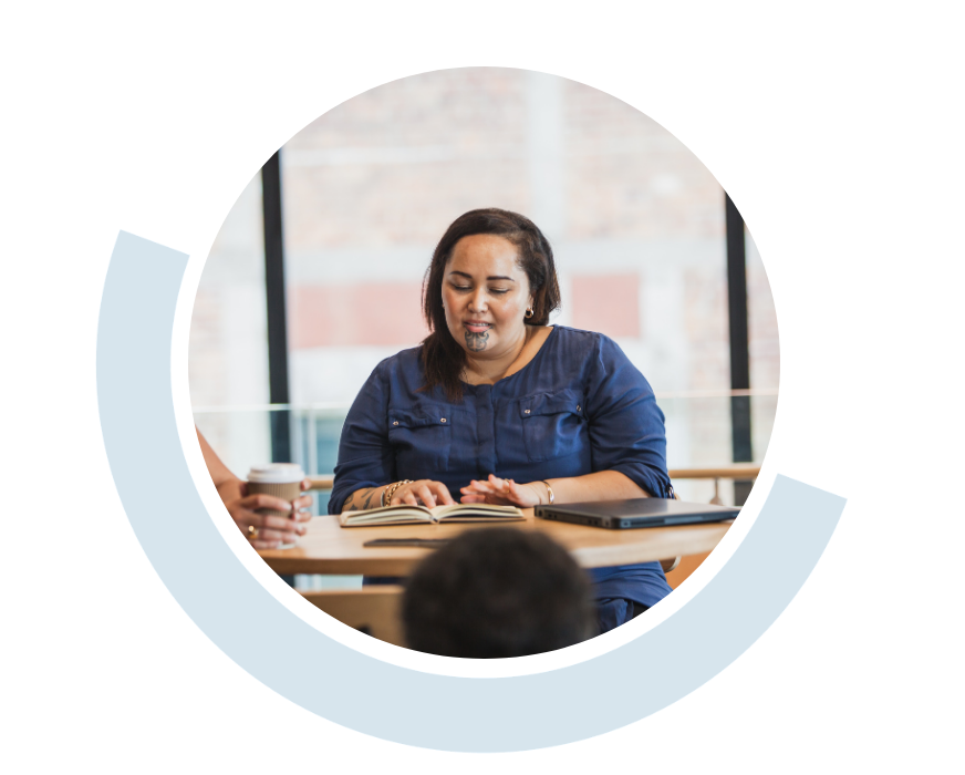 Website header graphic with a pale background and curved light blue accents, featuring a circular photo of a young Māori woman with a chin moko seated at a table speaking with a small group. Papers and other materials are spread across the table in front of her.
