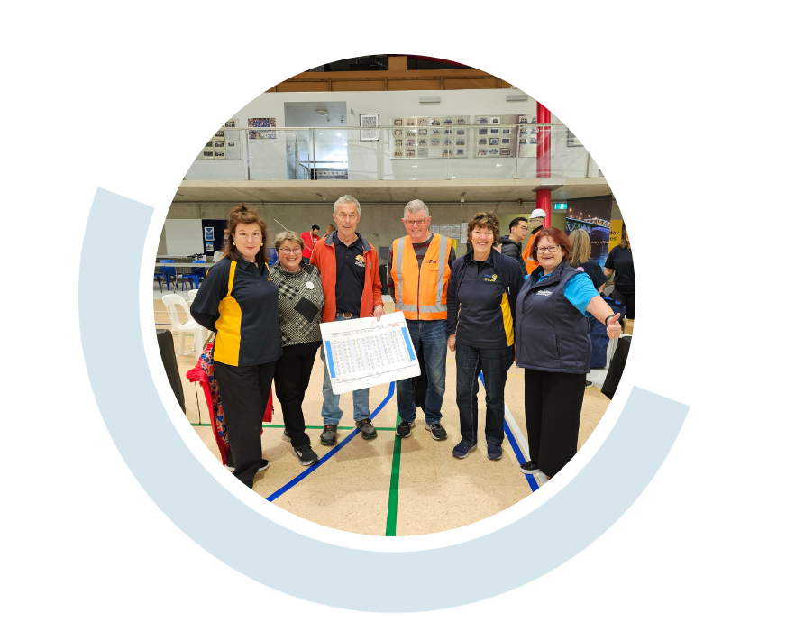 Website header graphic with a pale background and curved light blue accents, featuring a circular photo of a group of volunteers standing together indoors, one wearing bright orange high-visibility vests and smiling at the camera.