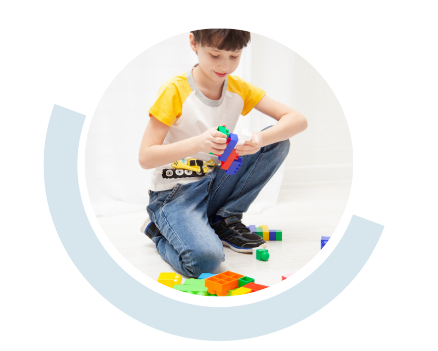 Website header graphic with a pale background and curved light blue accents, featuring a circular photo of a young boy sitting on the floor and playing with brightly coloured educational building blocks.