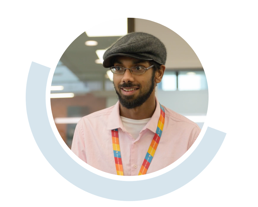 Website header graphic with a pale background and curved light blue accents, featuring a circular portrait of a smiling young man wearing glasses, a flat cap, a pale pink shirt and a colourful lanyard in a workplace setting.