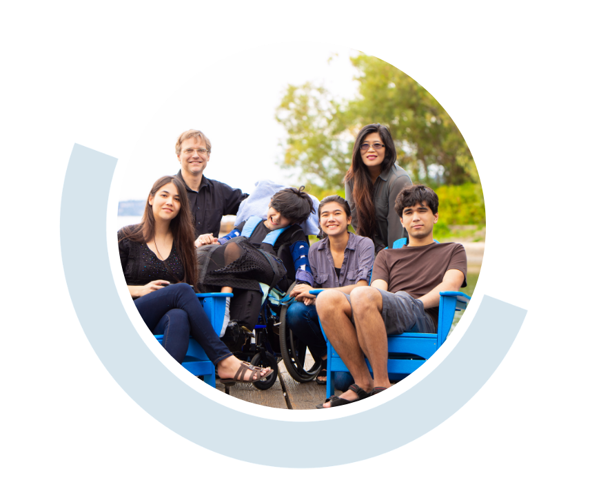 Website header graphic with a pale background and curved light blue accents, featuring a circular outdoor photo of a diverse group of adults and children gathered together and smiling, with a young person in a wheelchair in the centre of the photo.