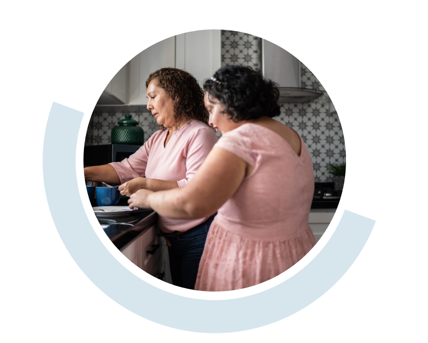 Website header graphic with a pale background and curved light blue accents, featuring a circular photo of two women preparing food together in a home kitchen, suggesting supported or shared living.