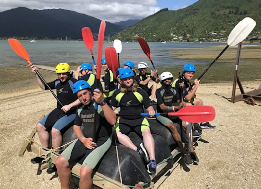 Outdoor group photo of nine people wearing wetsuits, life jackets and colourful helmets, sitting together on a large raft and raising kayak paddles in the air. They are gathered on a beach beside calm water, with green hills and mountains in the background.