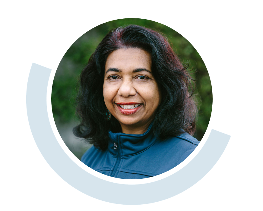 Website header graphic with a pale blue background and curved navy and bright blue accents, featuring a circular outdoor portrait of a smiling woman with shoulder-length dark hair and a blue jacket; counsellor Asoka Gamage-Mendes.