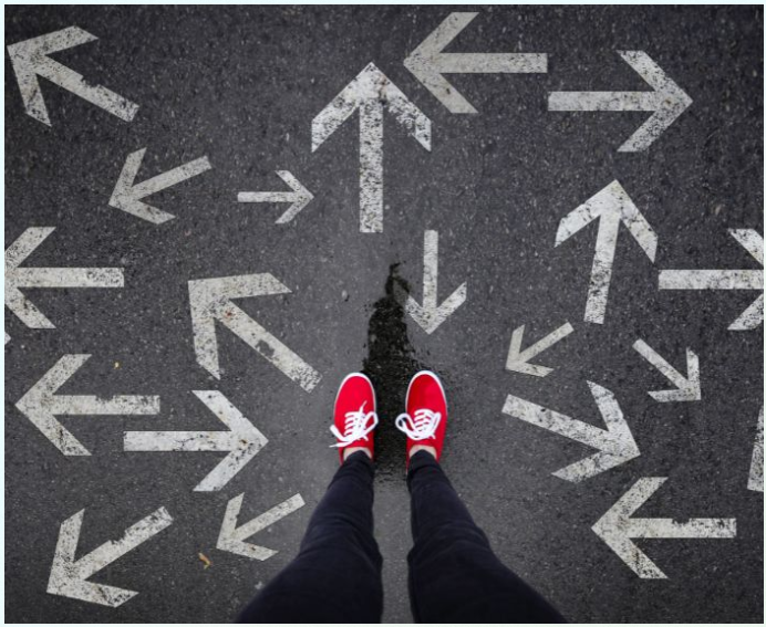 Overhead photo of a person in bright red shoes standing at the centre of many white arrows painted on dark pavement, pointing in different directions and symbolising choices, uncertainty or decision-making.